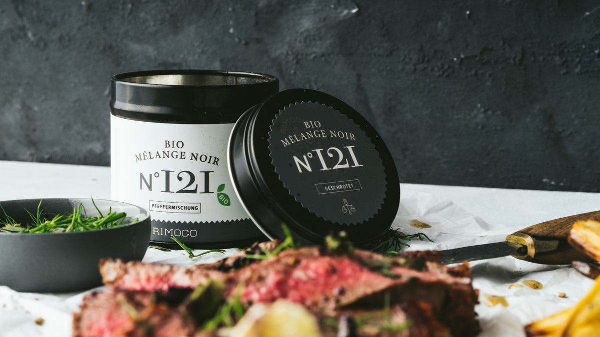 Aufgeschnittenes Fleisch und frische Kräuter neben einer Bio Mélange Noir N°12I-Gewürzdose auf einem Holztisch.
