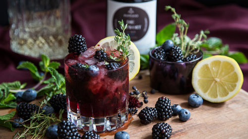 Zwei Beerencocktails mit Zitrone und Kräutern neben Geschenkset mit Gin und würzigen Botanicals.