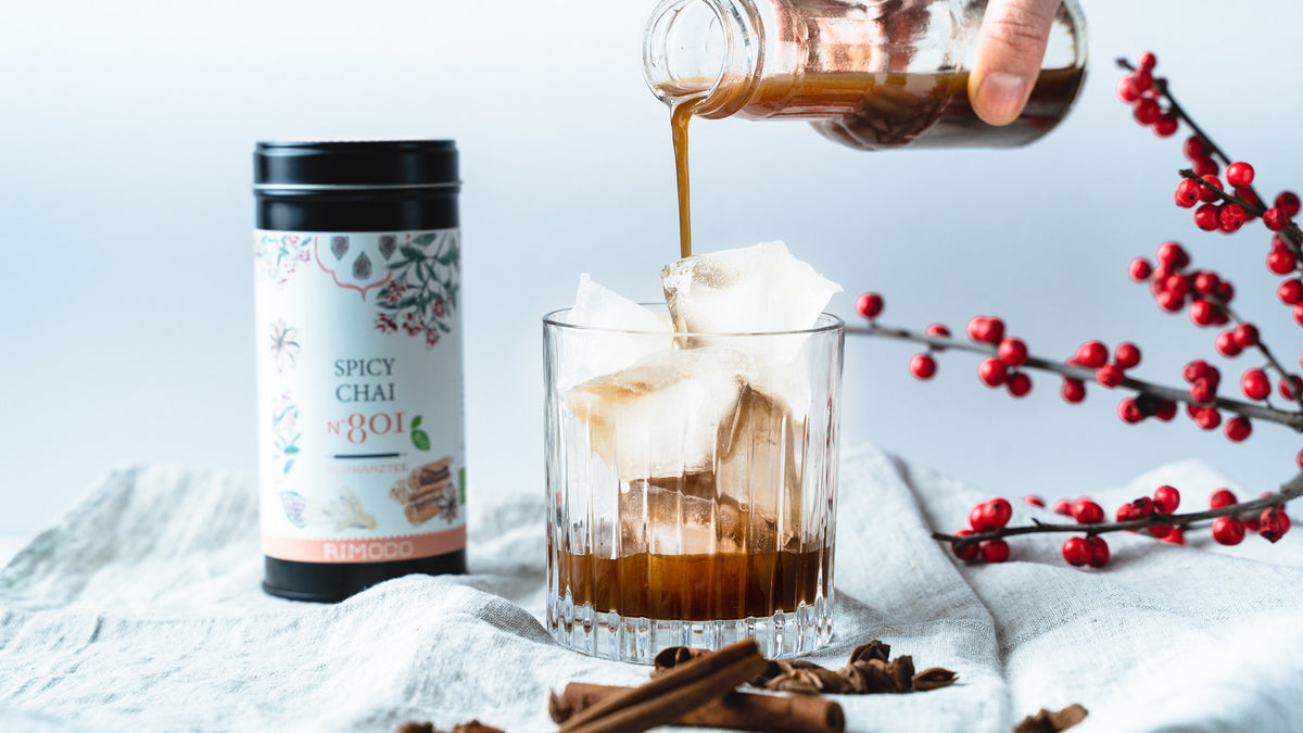 Eistee-Chai im Glas mit Eis, daneben eine Chai-Dose und rote Beeren auf einem weichen Tuch.