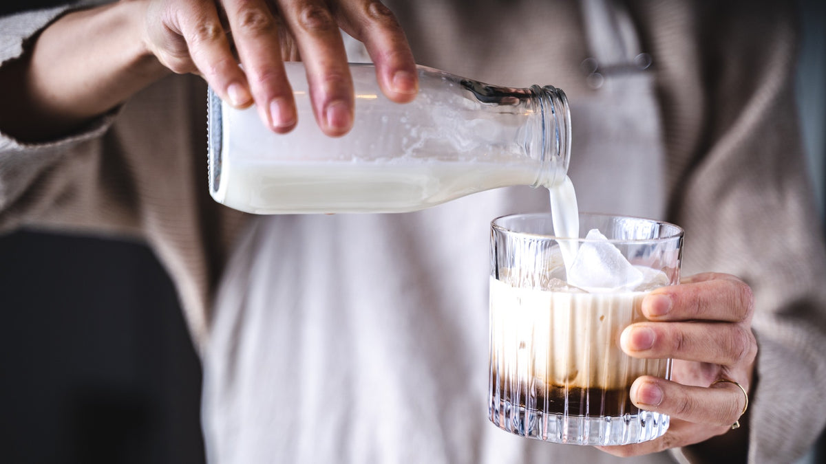 Milch wird aus einer Glasflasche in einen Eiskaffee gegossen und bildet dabei eine cremige Wirbeloptik.