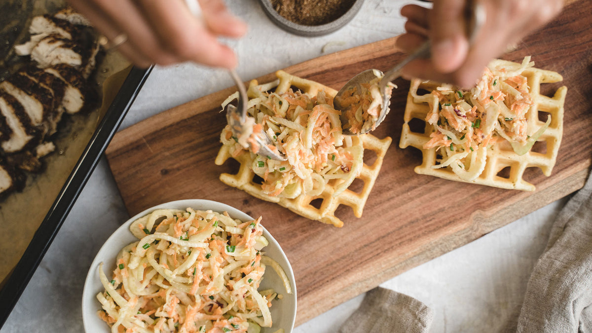 Auf einem Holzbrett liegen Waffeln mit frischem Gemüseslaw, daneben steht eine Schüssel mit Slaw.