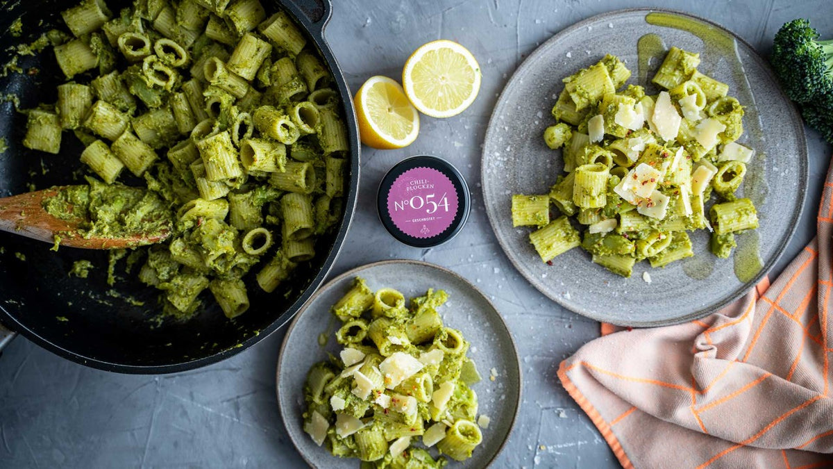 Pesto-Pasta mit Zitrone und Parmesan auf zwei Tellern und in einer Pfanne, rosa Etikett auf einem grauen Tisch.