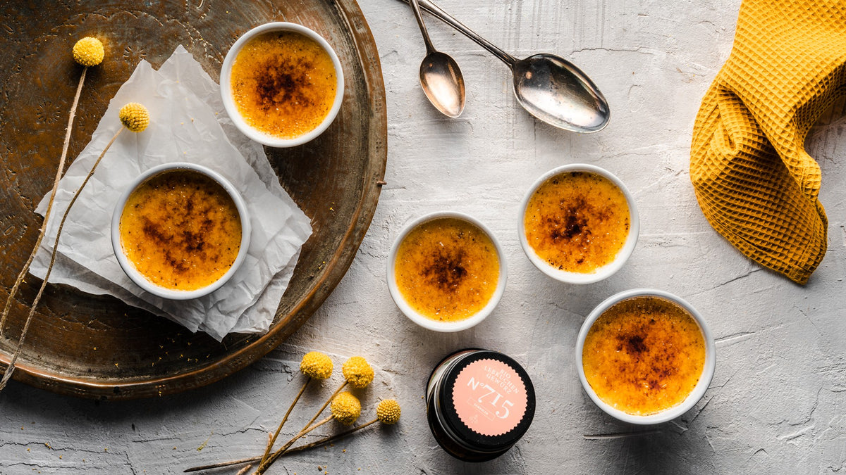 Fünf Schälchen Crème Brûlée auf einem Tisch, dazu Löffel, ein Metalltablett und ein gelbes Tuch.