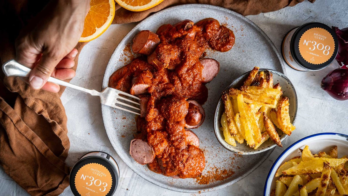 Eine Hand spießt eine Wurst mit roter Soße auf einem Teller neben Pommes und zwei Gewürzgläsern auf.