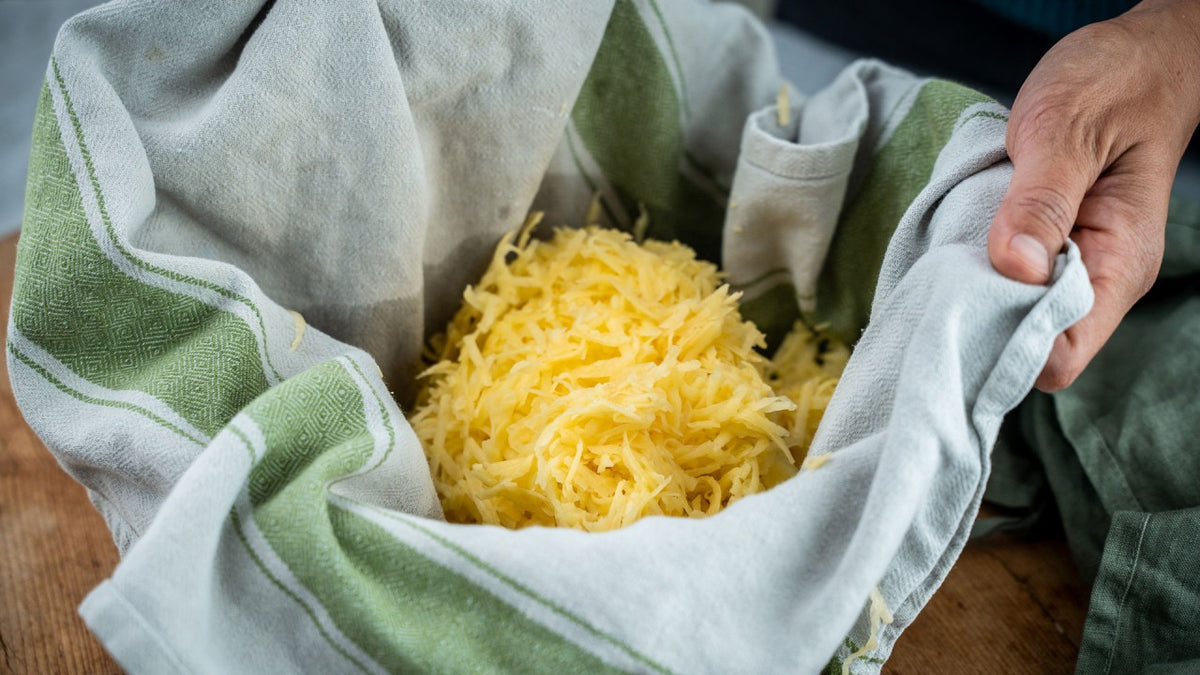 Geriebene Kartoffeln in einer mit einem Tuch ausgelegten Schüssel, wobei die Hände die Tuchränder festhalten.