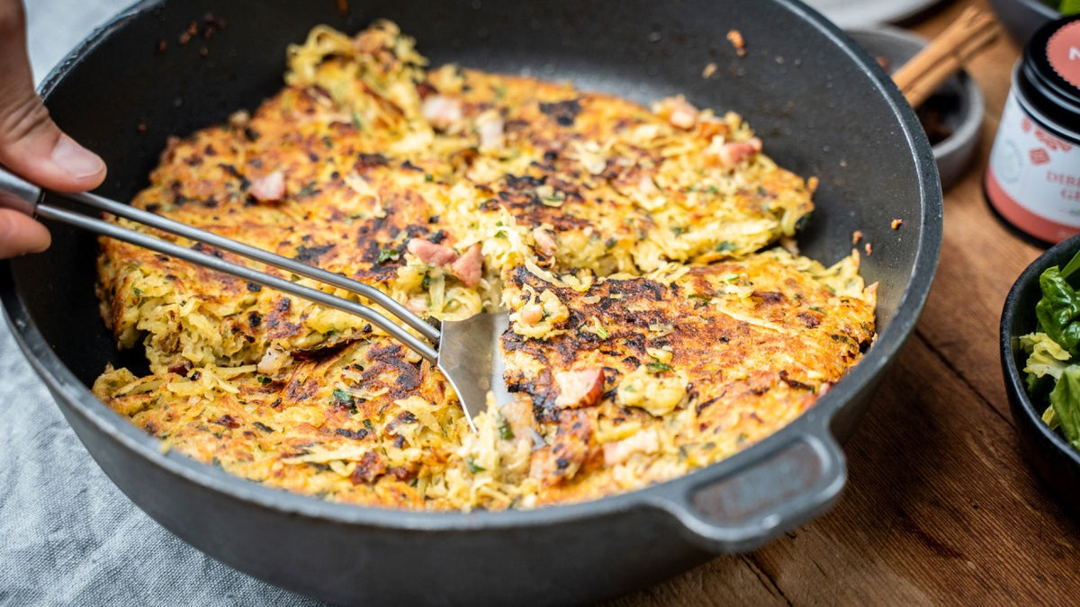 Eine Hand schneidet einen goldbraunen, knusprigen Kartoffelrösti mit Schinken in einer Pfanne auf einem Holztisch auf.
