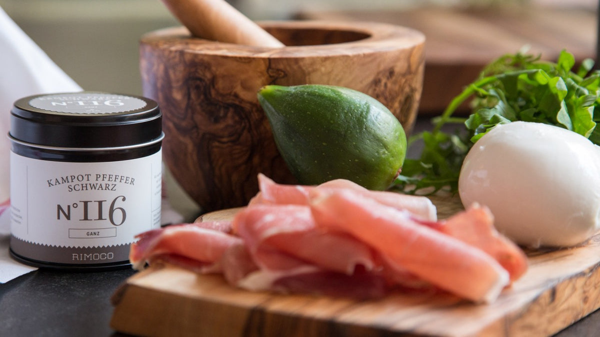 Prosciutto, Mozzarella, grüne Papaya, grober schwarzer Pfeffer und Kräuter, auf einem Holzbrett angerichtet.