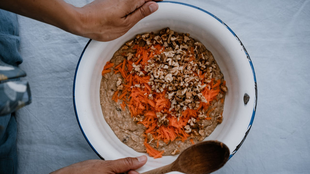 Eine Schüssel mit Karottenkuchenteig, geraspelten Karotten und Nüssen, die von Händen und einem Holzlöffel gehalten wird.