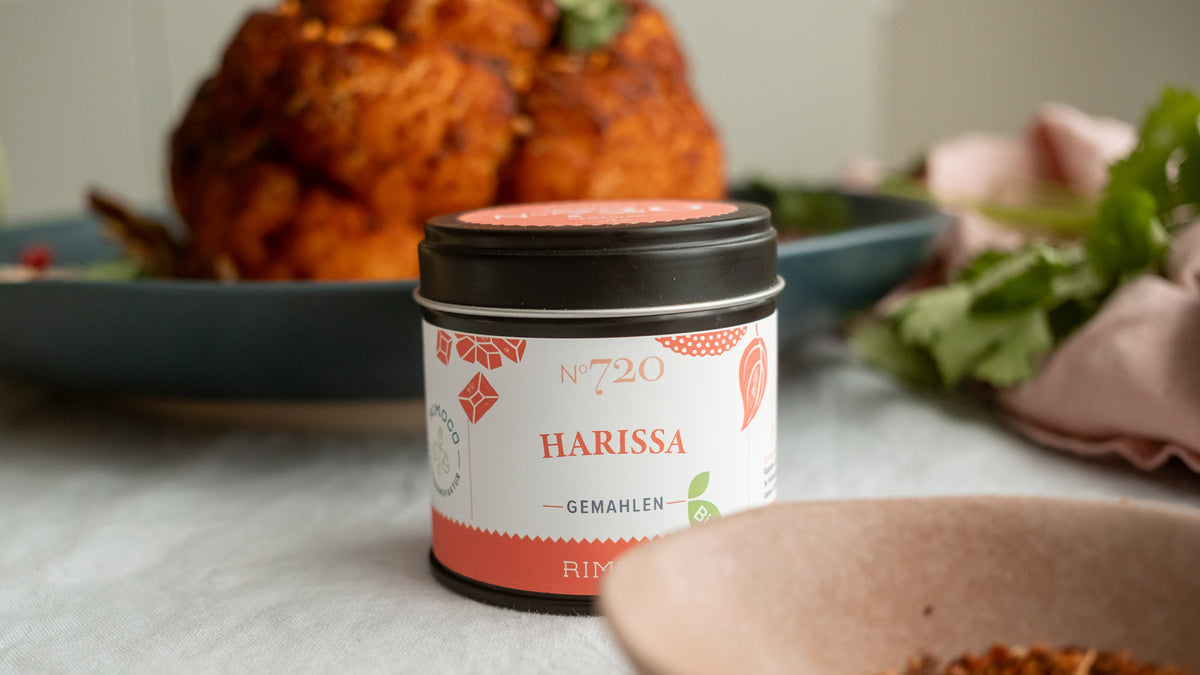Auf einem Tisch steht eine Dose gemahlene Harissa, umgeben von frischen Kräutern und Lebensmitteln.