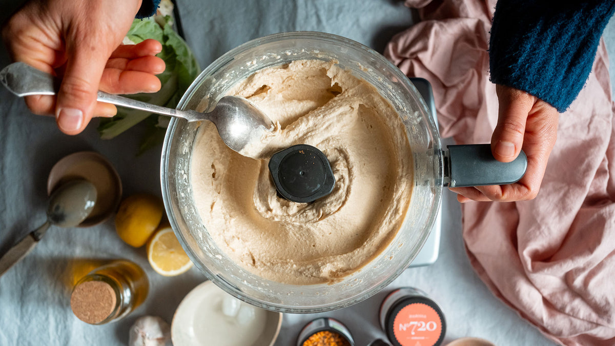 Ein Mixer mit cremigem Hummus wird in den Händen gehalten, daneben liegen frische Kichererbsen und Knoblauch.