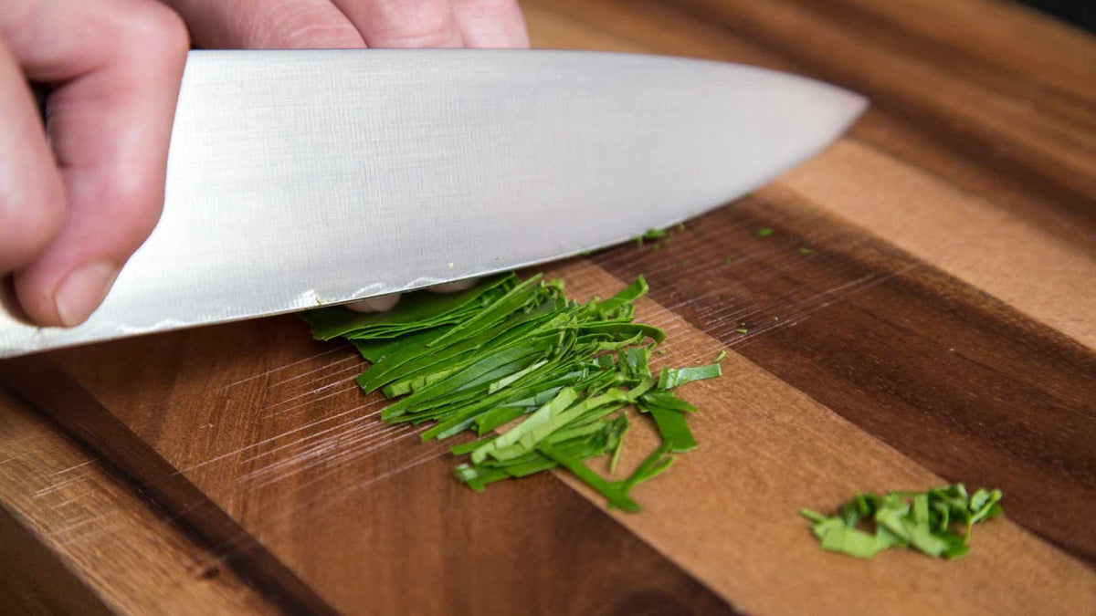 Hand schneidet frische grüne Kräuter mit einem großen Messer auf einem Holzbrett in feine Stücke.