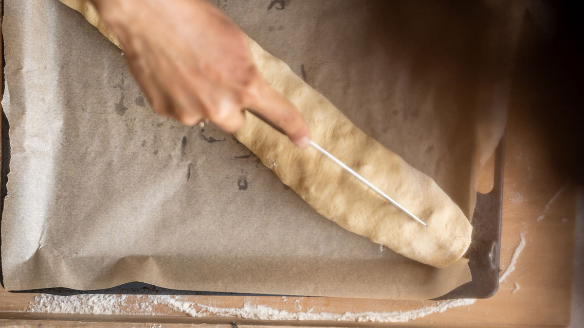 Eine Hand schneidet mit einem Messer einen rustikalen Brotteig auf einem mit Backpapier ausgelegten Backblech ein.