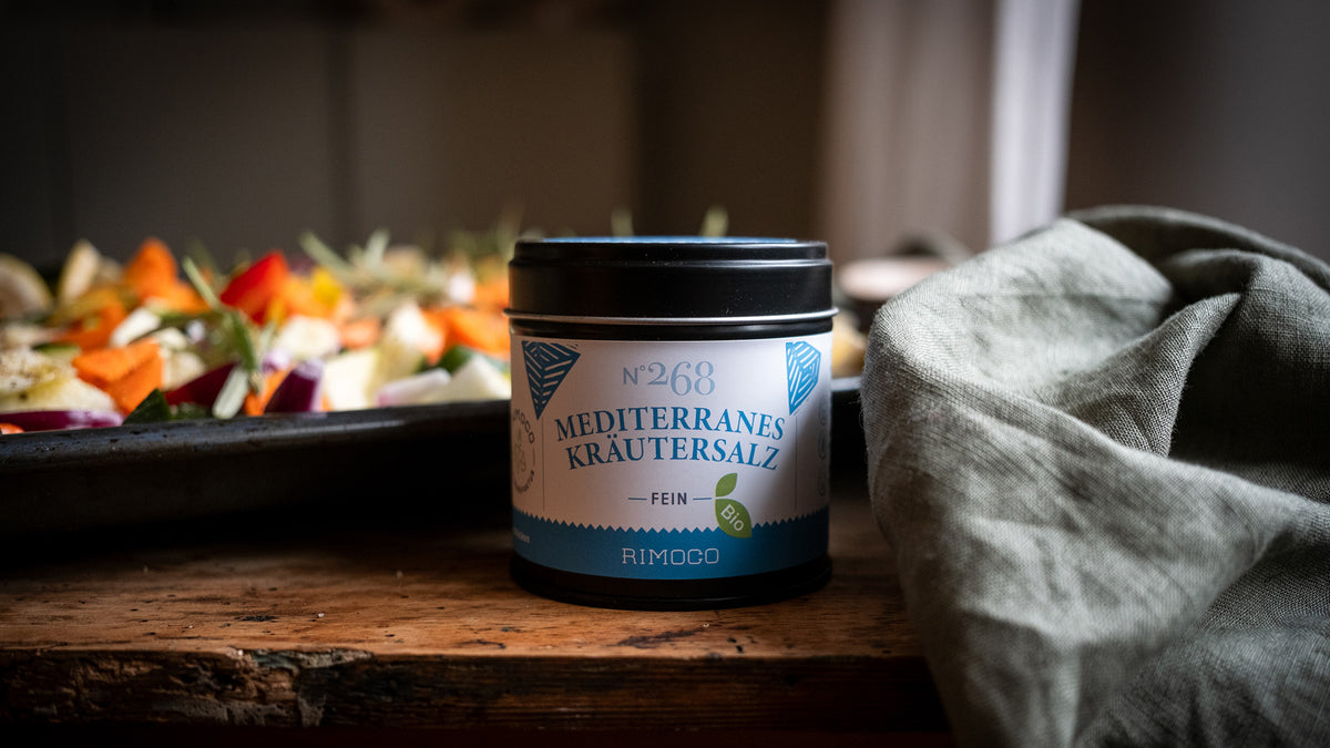 Mediterranes Kräutersalz in einer Dose auf einem Holztisch neben einem Backblech und einem grünen Tuch.