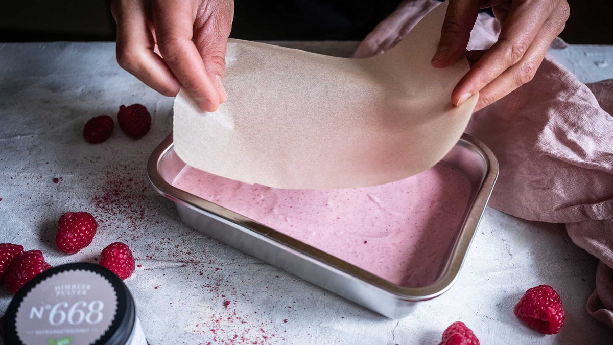 Hände bedecken eine Form mit einer rosa Himbeermischung, daneben liegen frische Himbeeren.