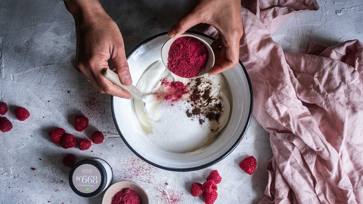 Geben Sie Himbeerpulver zu Joghurt in einer Schüssel, legen Sie frische Himbeeren daneben und legen Sie ein rosa Tuch daneben.