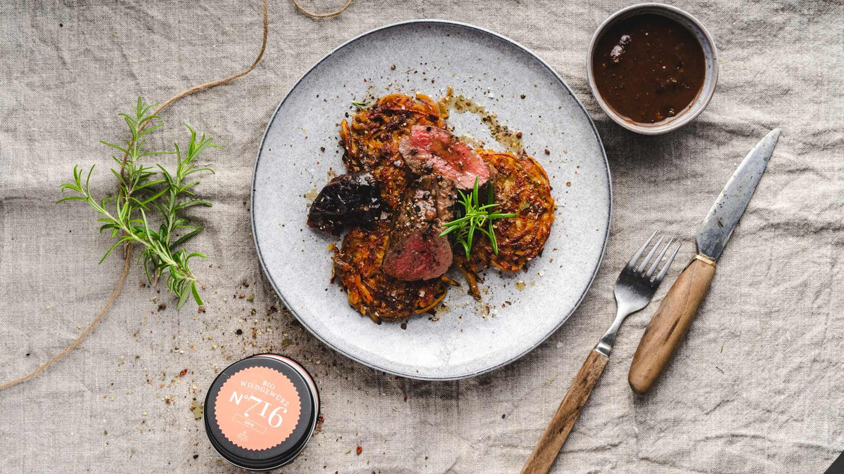 Ein Teller mit Steak, Rösti, Kräutergarnitur, Sauce, Besteck und Gewürzen auf einer Leinentischdecke.