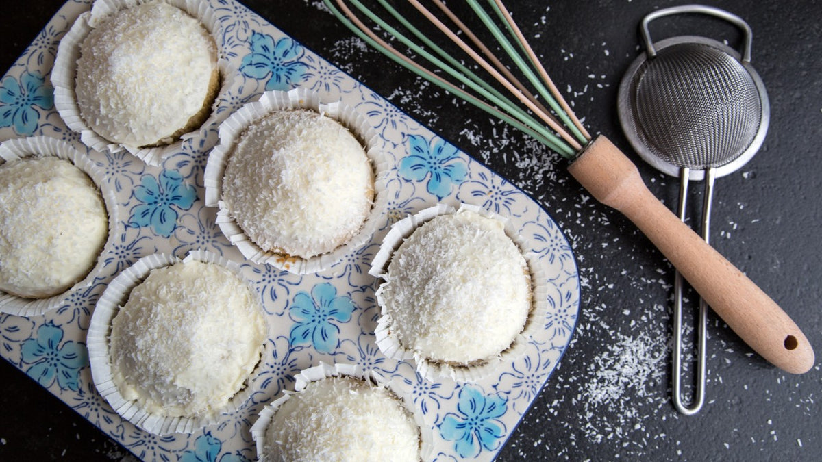 Sechs mit Kokosraspeln bestreute Cupcakes auf einem Tablett mit Blumenmuster, daneben ein Schneebesen und ein Sieb auf schwarzem Hintergrund.
