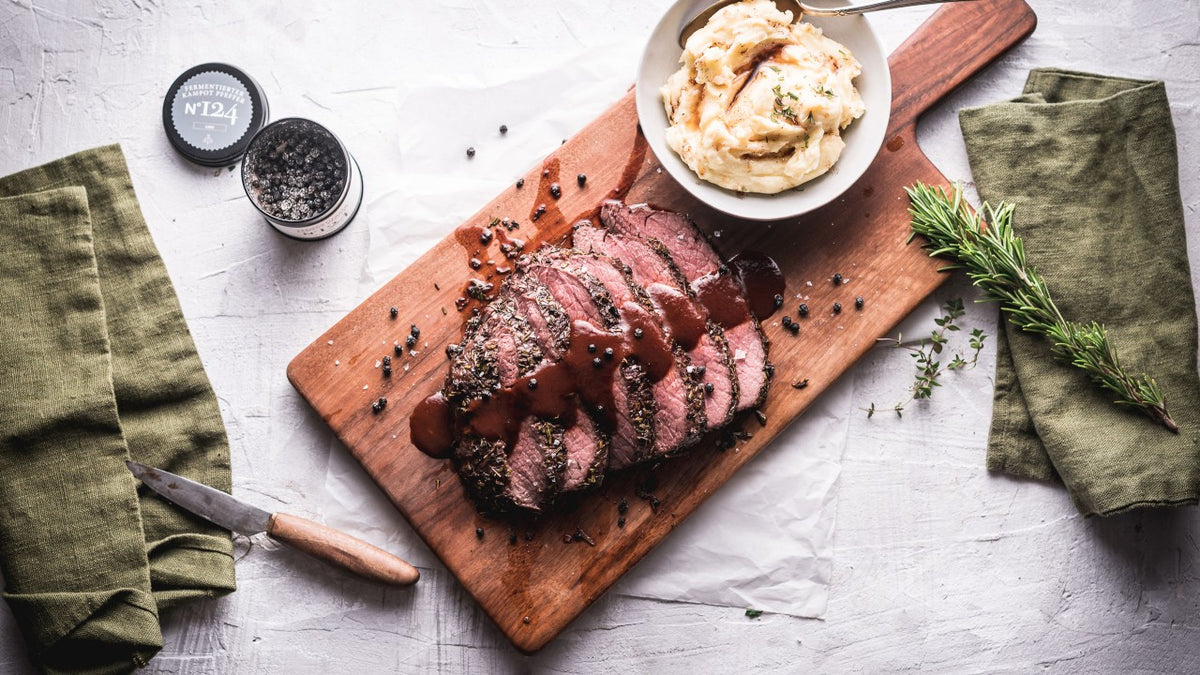 Roastbeefscheiben mit Soße, Kartoffelpüree, frischen Kräutern und Gewürzen auf einem Holzbrett.