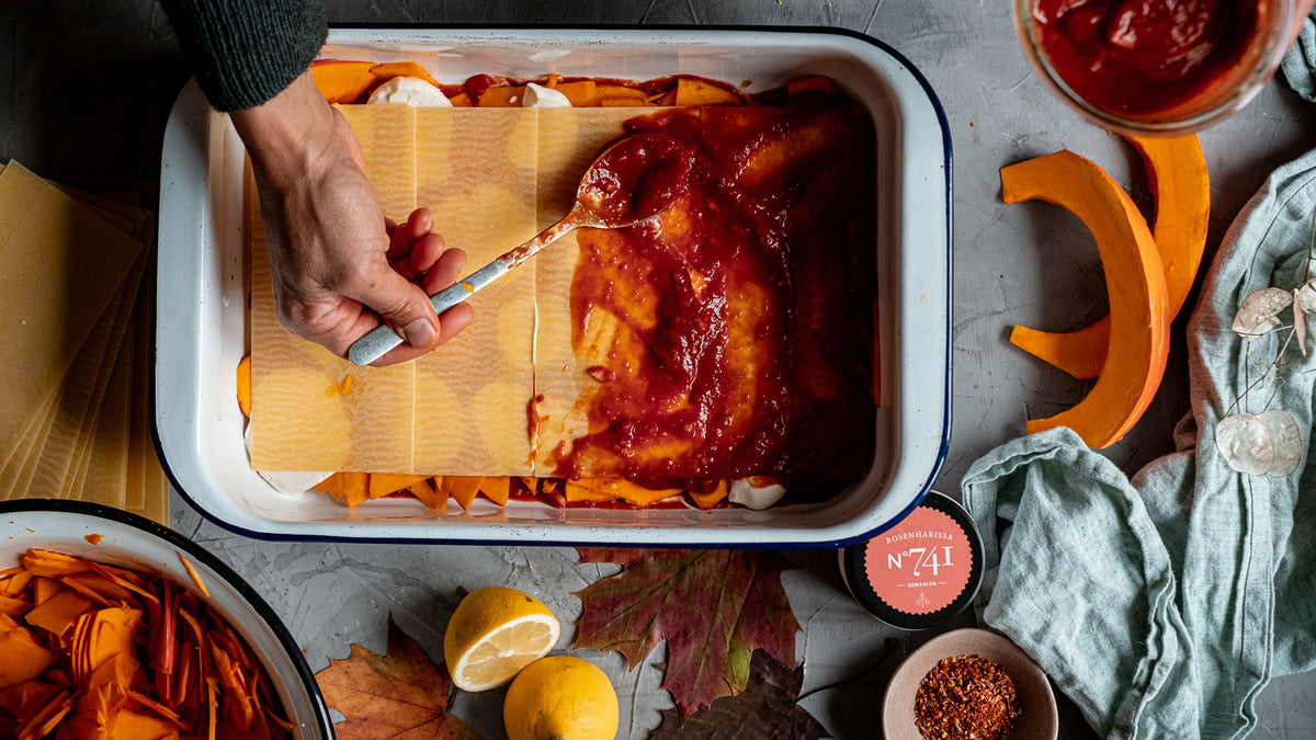 Lasagne in einer Auflaufform, über die mit der Hand Tomatensauce verteilt wurde, umgeben von herbstlichen Zutaten.
