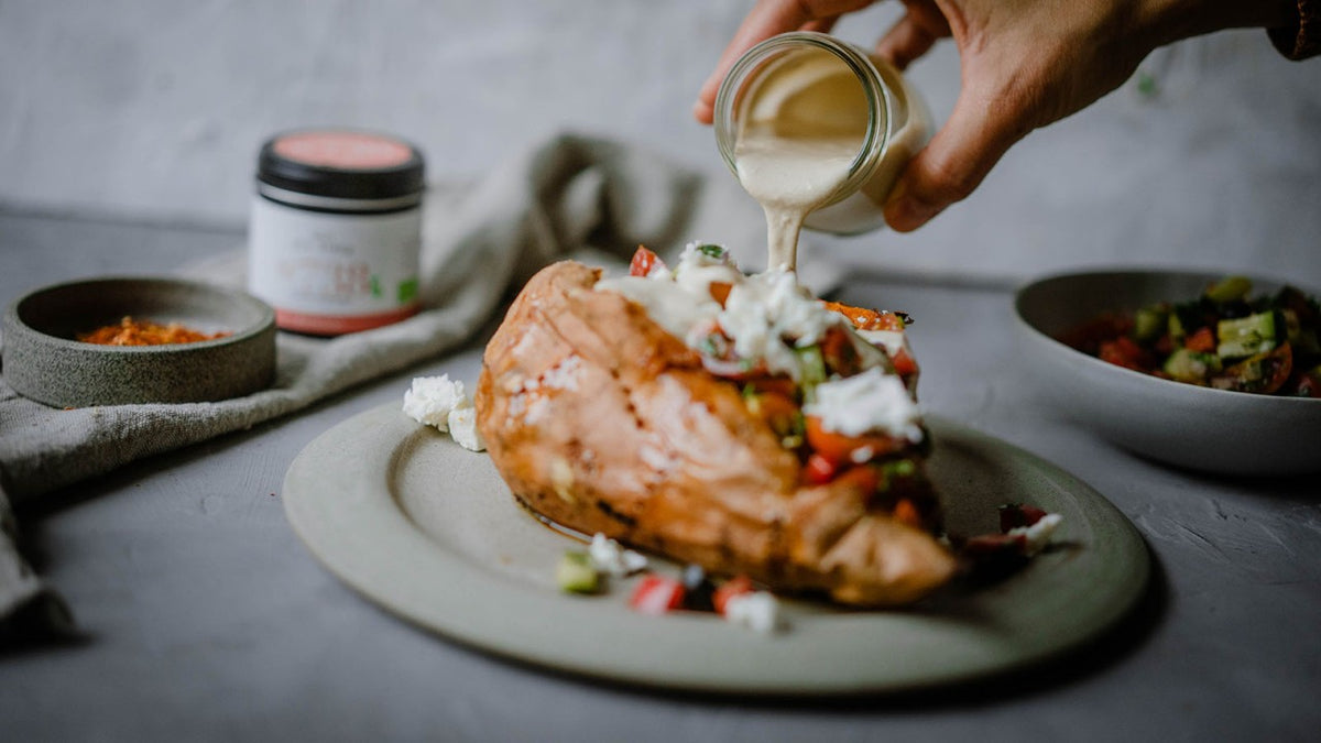 Eine Hand gießt eine Sahnesauce über eine gebackene Süßkartoffel mit Gemüse und Käse.