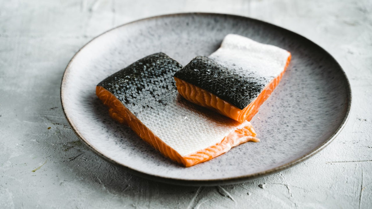 Zwei frische Lachsfilets mit Haut liegen auf einem grauen Keramikteller vor hellem Hintergrund.