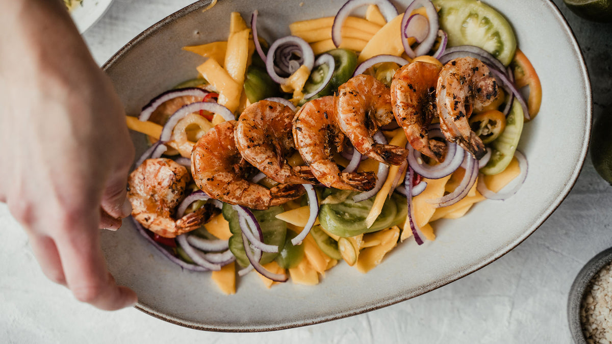 Gegrillte Garnelen auf Mango, Tomaten und Zwiebeln auf Teller, daneben Grillgewürze Geschenkbox N°1.