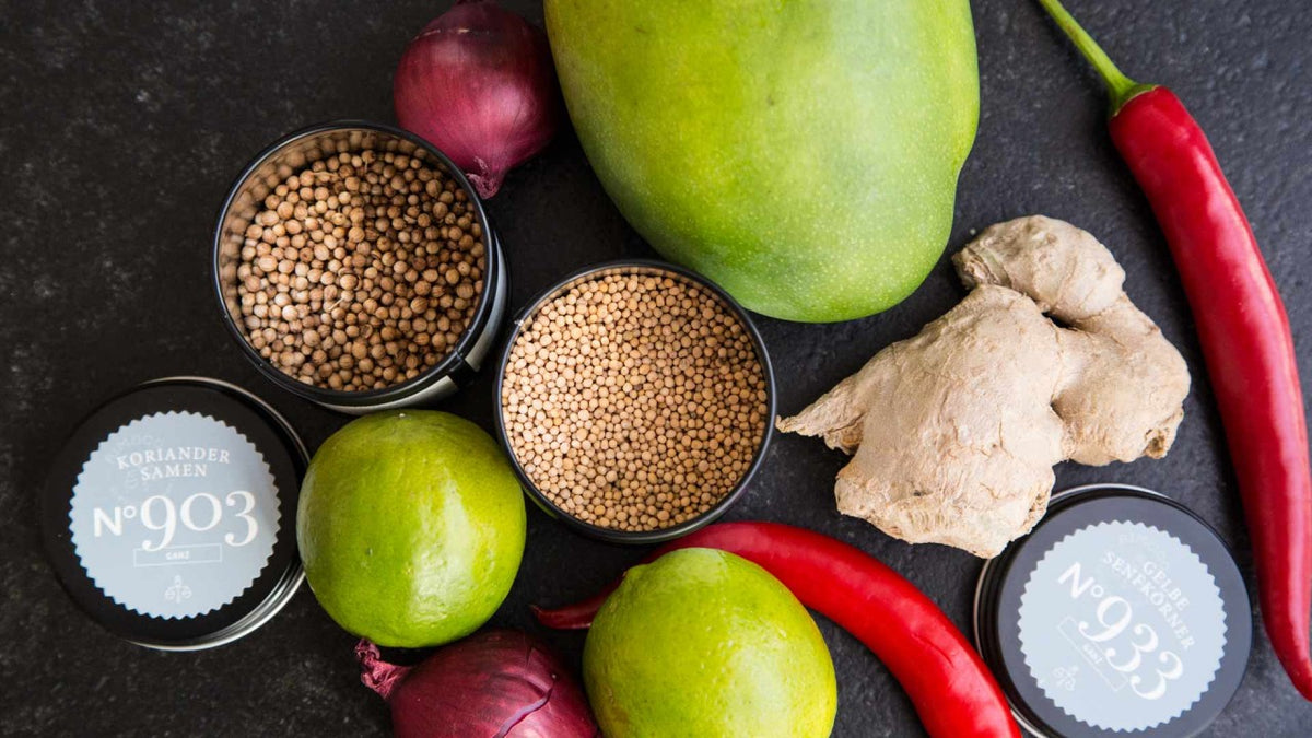 Limetten, Mango, Ingwer, rote Chilischoten, Zwiebeln und Koriandersamen vor dunklem Hintergrund.