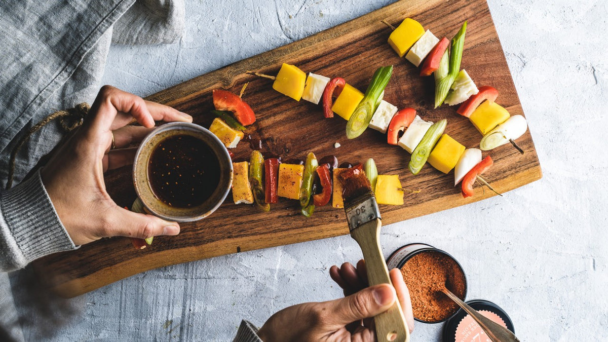 Mit den Händen die Gemüsespieße auf einem Brett mit Soße bestreichen, dazu Paprika, Zwiebeln und Mango.