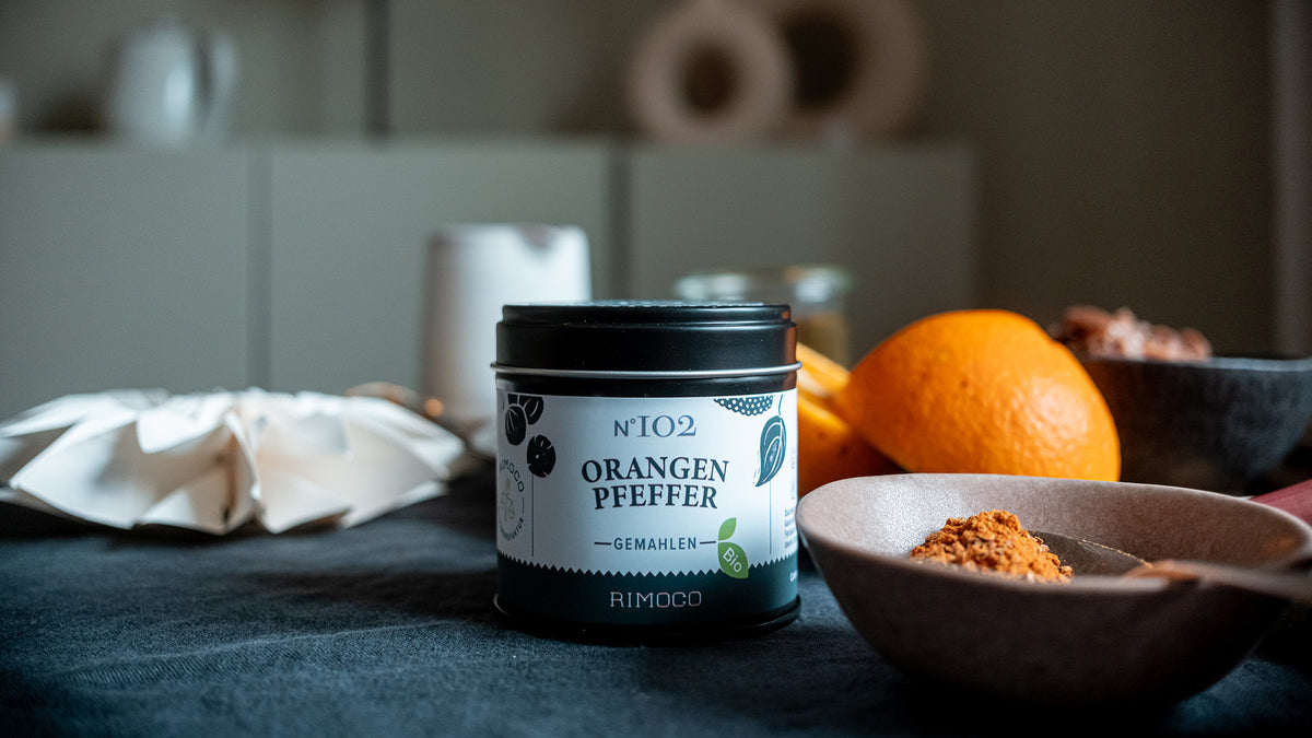 Eine Dose mit gemahlenem Orangenpfeffer auf dem Tisch, daneben frische Orangen und die leuchtend gelbe Schale des Gewürzes.