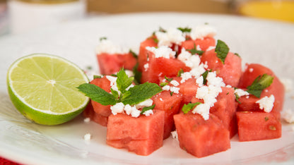 Ein Teller mit Wassermelonenwürfeln, Feta, Minze, Mango, Chili und Honig, dazu eine halbe Limette.