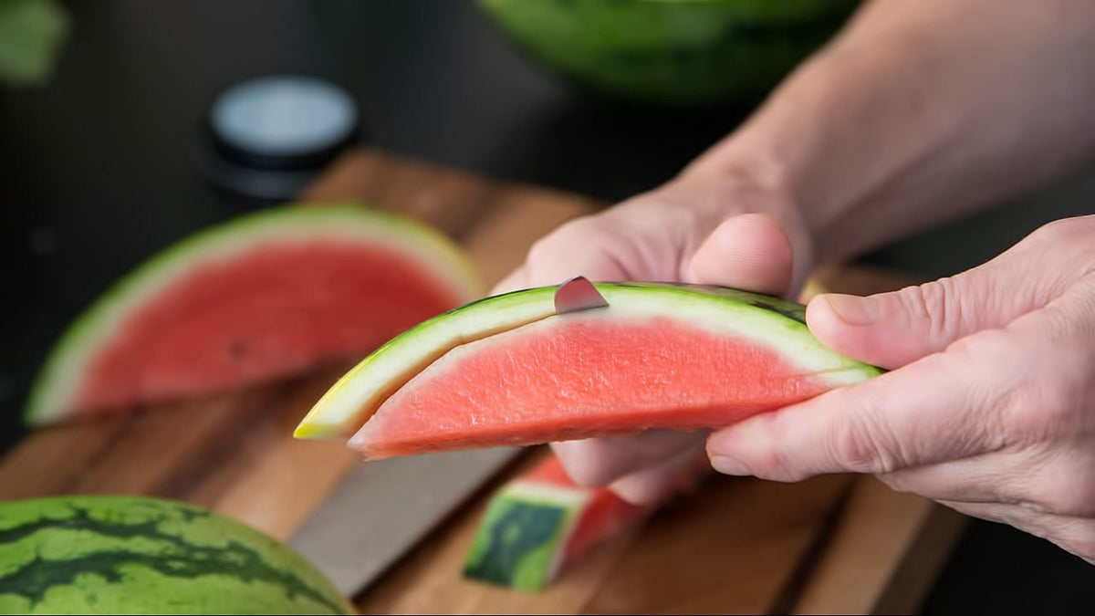 Eine Person schält mit einem kleinen Messer die grüne Schale von einer Wassermelonenscheibe auf einem Holzbrett.