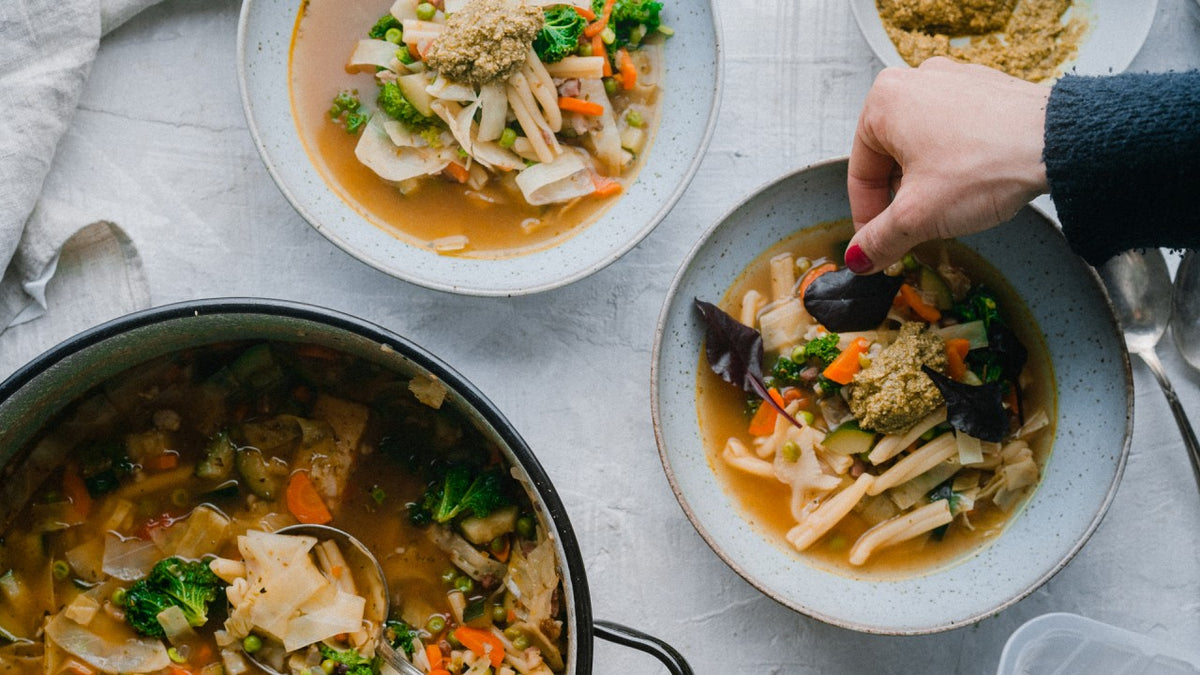 Zwei Schüsseln dampfende Gemüsenudelsuppe, eine davon frisch von Hand garniert, daneben ein Topf.