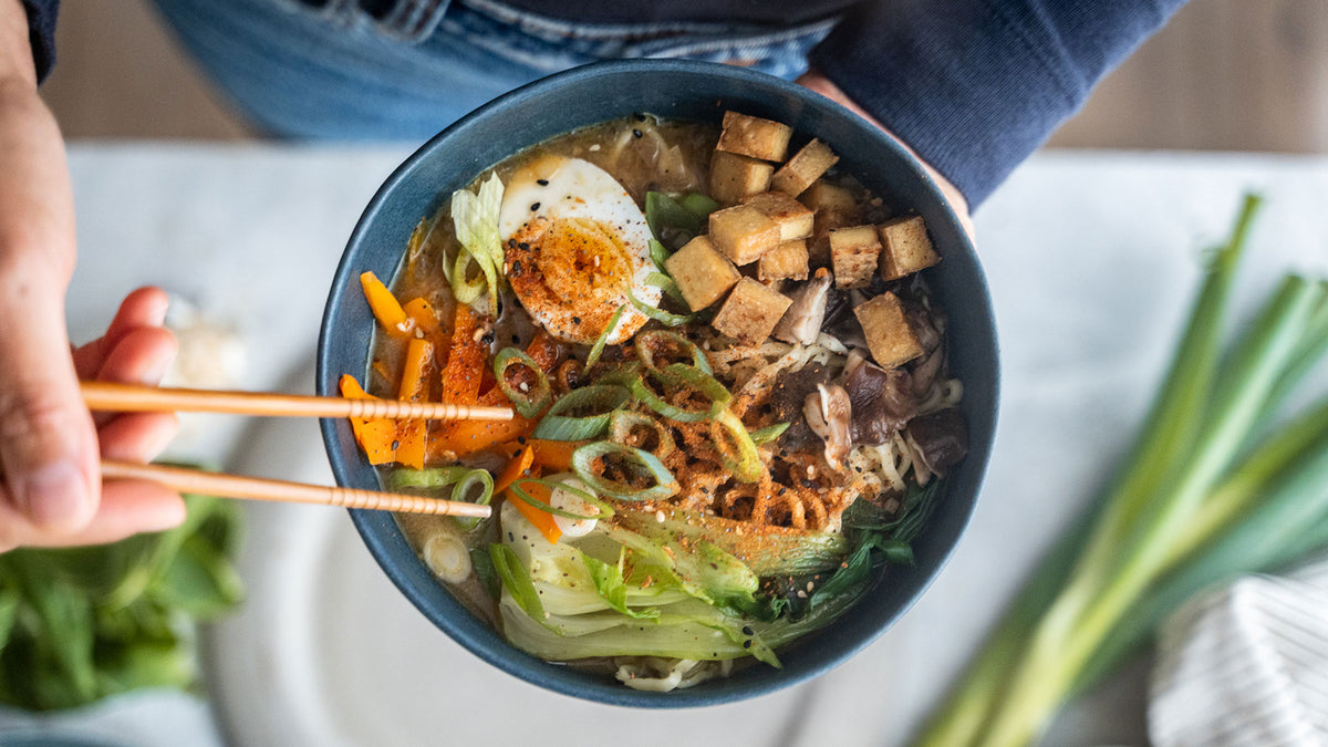 Eine Person hält eine Schüssel Ramen mit Tofu, Ei, Gemüse und Essstäbchen in der Hand.
