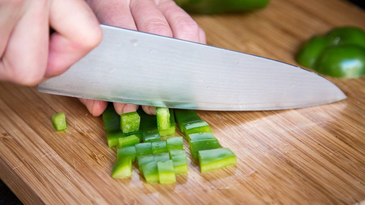 Eine Hand schneidet mit einem großen Messer grüne Paprika auf einem Holzschneidebrett in Würfel.
