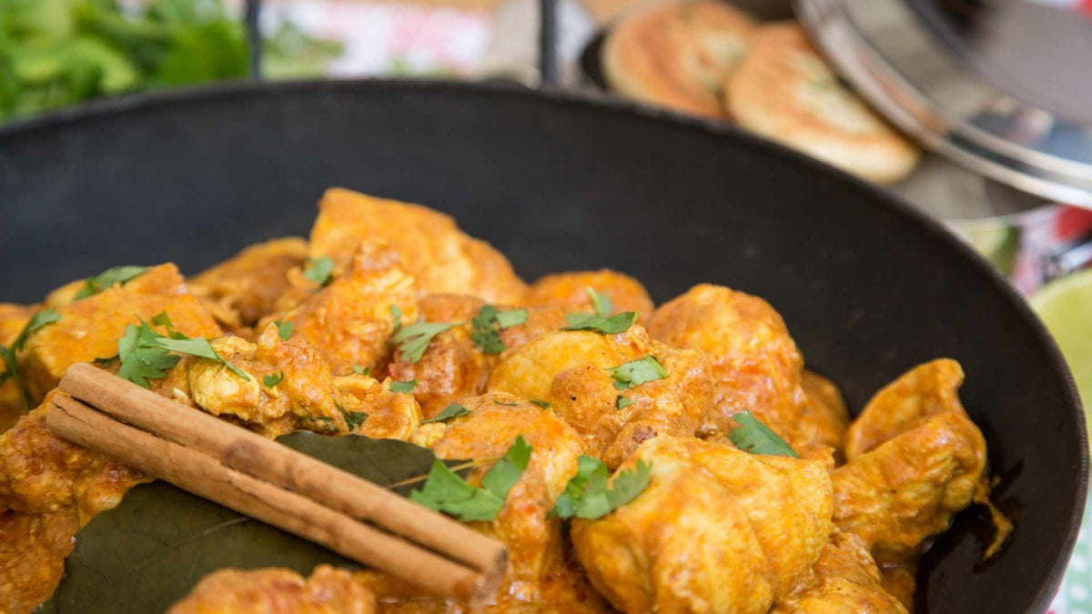 Nahaufnahme von Hähnchencurry mit Koriander, Lorbeerblättern und Zimtstangen in einer schwarzen Schüssel.