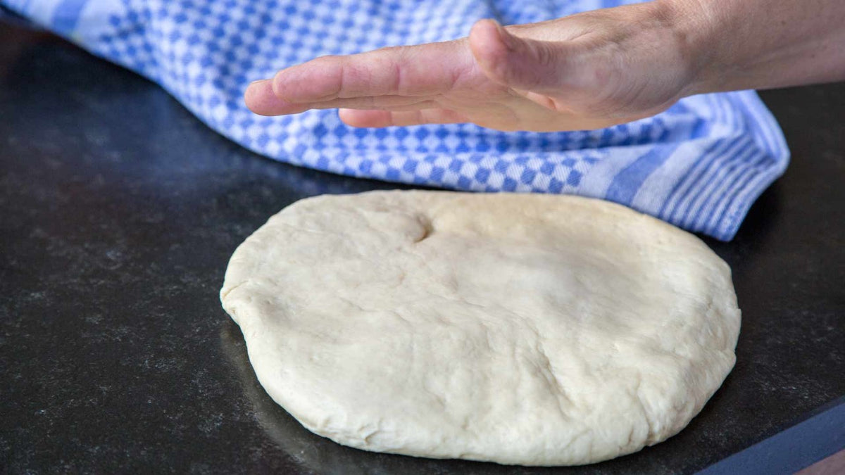 Eine Hand schwebt über Pizzateig auf einer dunklen Arbeitsfläche neben einem blau karierten Tuch.