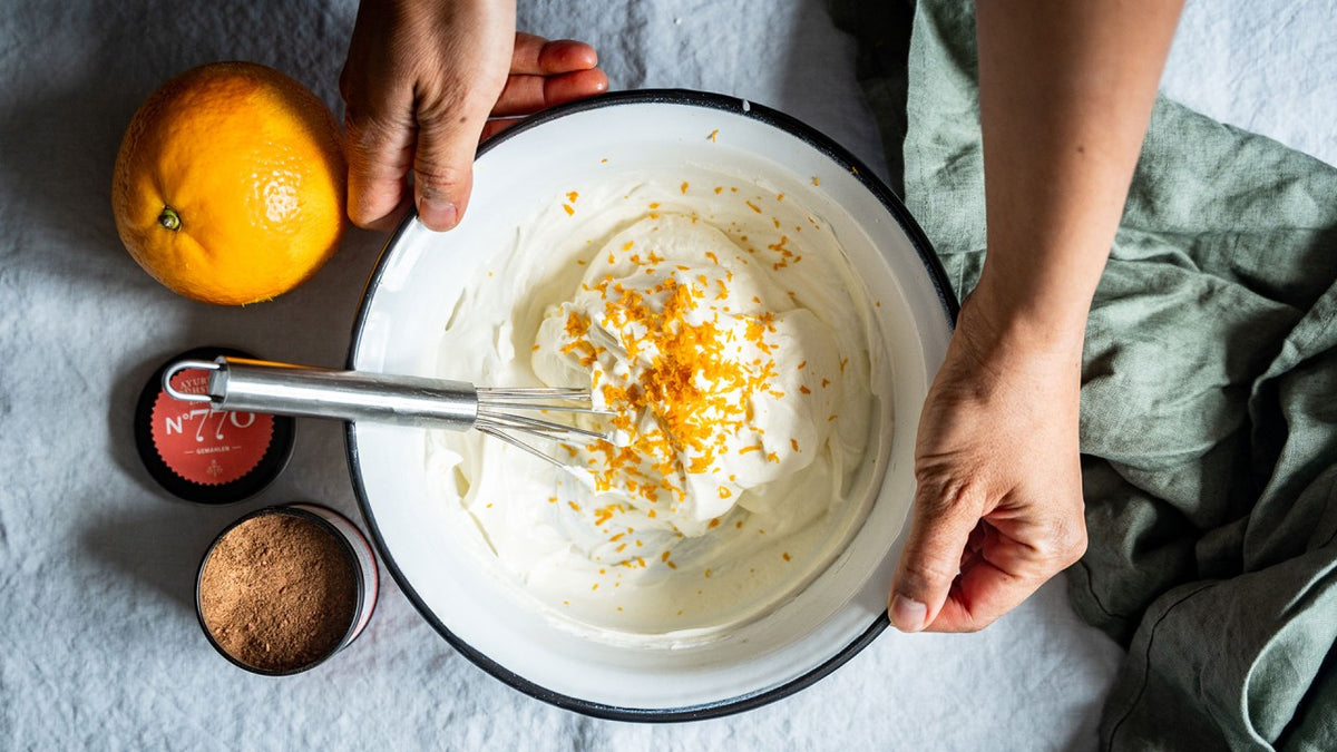 In den Händen wird eine Schüssel mit Schlagsahne, Orangenschale und Kakaopulver gehalten, daneben liegt ein Schneebesen.
