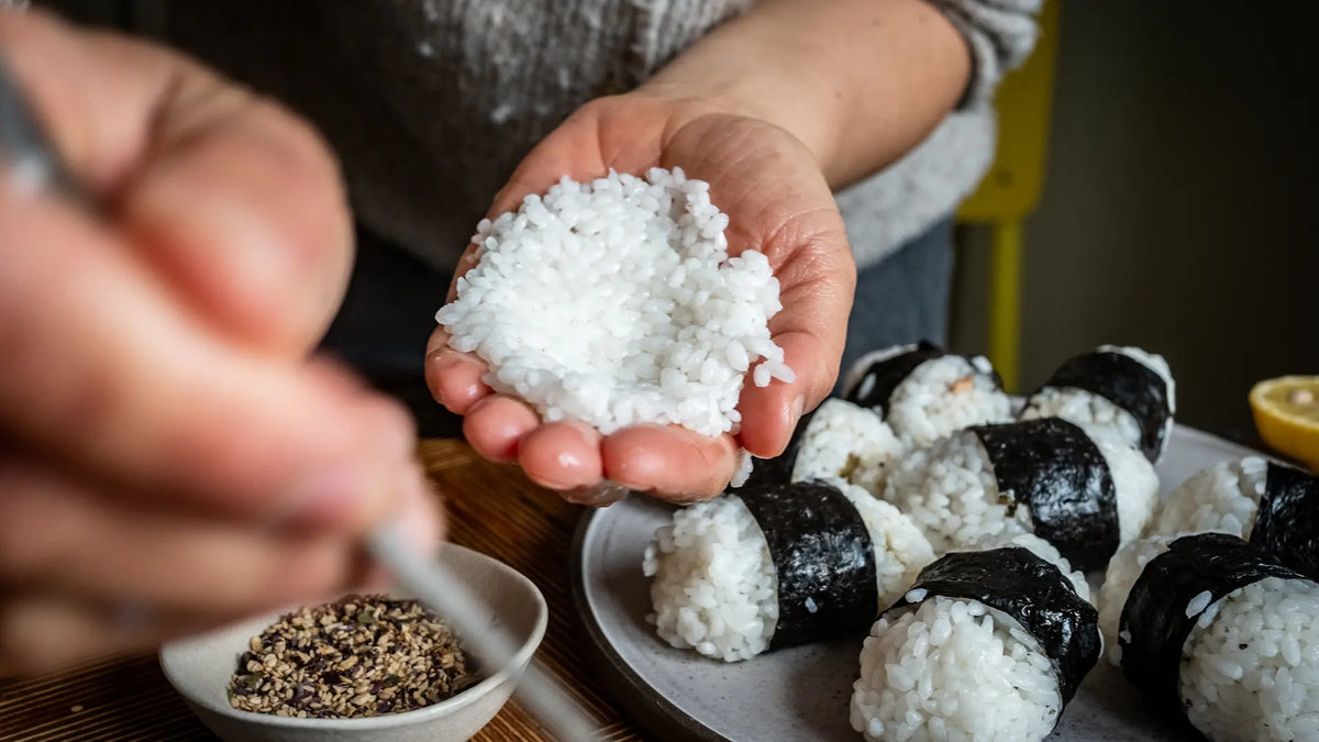 Hände formen Reis für Onigiri, im Hintergrund fertige Stücke und sichtbare Gewürze.