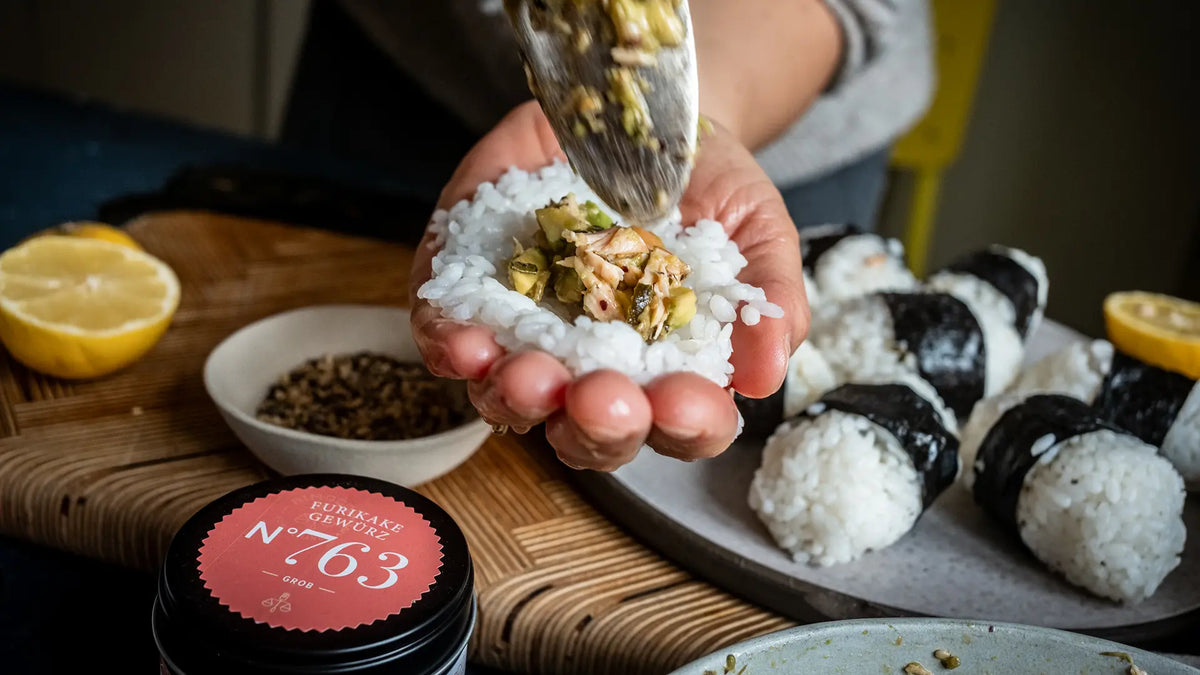 Ein Mensch füllt Onigiri mit einer Mischung, daneben liegen frisches Sushi und weitere Onigiri.