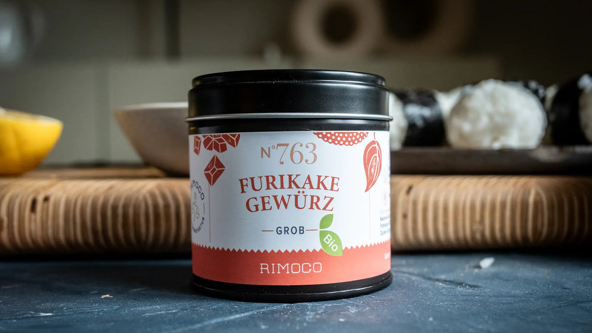 Eine Rimoco-Furikake-Gewürzdose steht auf einem Tisch, im Hintergrund sind Teller und Essen unscharf zu sehen.
