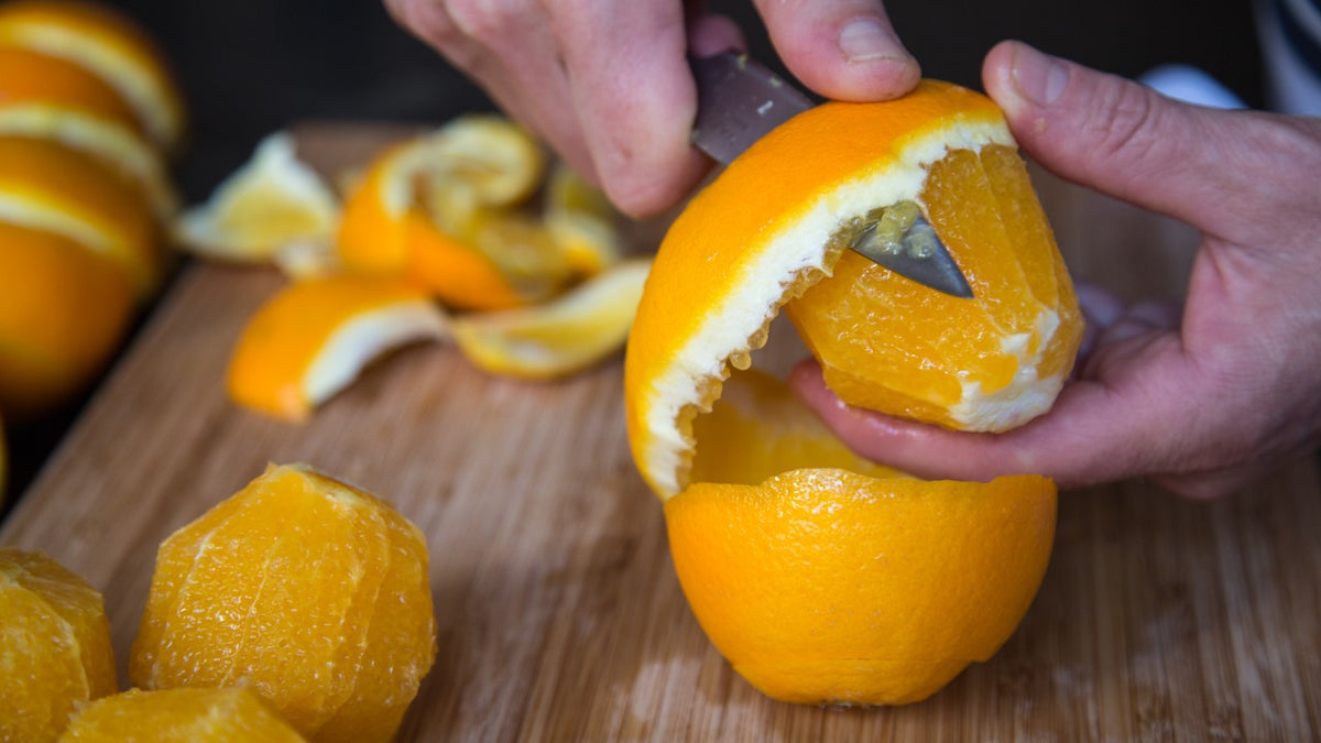 Eine Person schneidet eine Orange auf einem Holzbrett; daneben liegen geschälte Orangen.