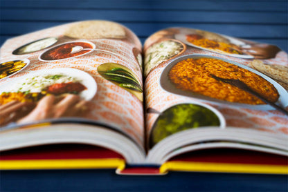 Geöffnetes Original Indisch-Kochbuch mit Fotos von buntem vegetarischem Curry auf dunkelblauem Tisch.