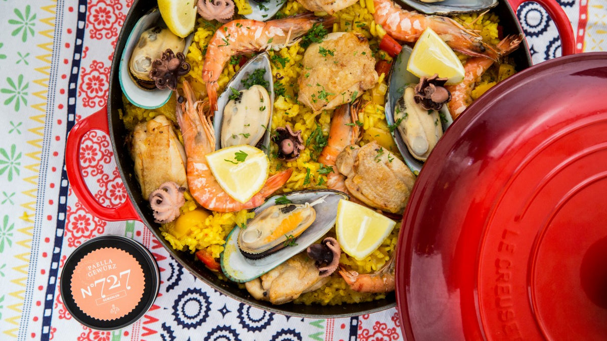Spanische Meeresfrüchte-Paella mit Garnelen, Muscheln, Tintenfisch, Zitronenschnitzen und Reis neben einem roten Deckel.