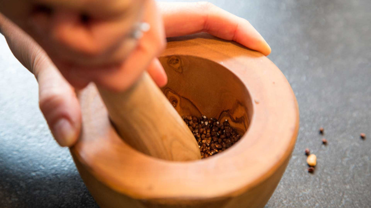 Eine Hand zermahlt Gewürze mit einem Holzstößel im Mörser. Grobe Gewürzstücke sind sichtbar.