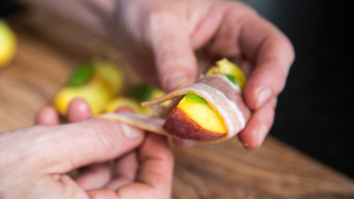 Auf rustikalem Holz wickeln Hände eine Pfirsichscheibe und ein grünes Blatt mit Prosciutto ein.