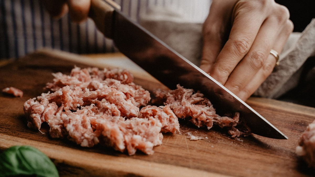 Jemand hackt rohes Hackfleisch mit einem großen Messer auf einem Holzbrett klein.