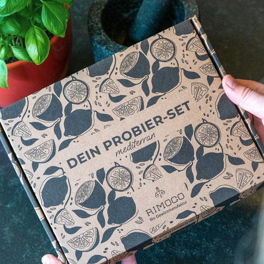 Hände halten eine Probierset Mediterrane Gewürze Box mit Aufschrift DEIN PROBIER-SET RIMOCO neben einer Pflanze.