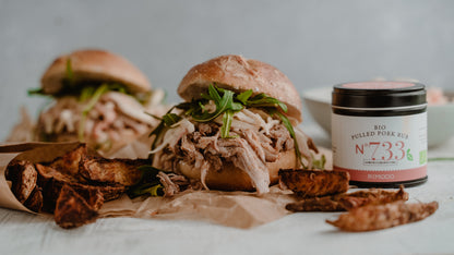 Pulled Pork Sandwiches mit Salat, Kartoffelecken und einer BBQ Rubs Geschenkbox auf rustikalem Untergrund.