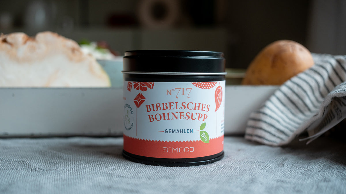 Eine Dose „Bibbelsches Bohnesupp“ auf einem Tisch neben rustikalem Brot und einem gestreiften Tuch.