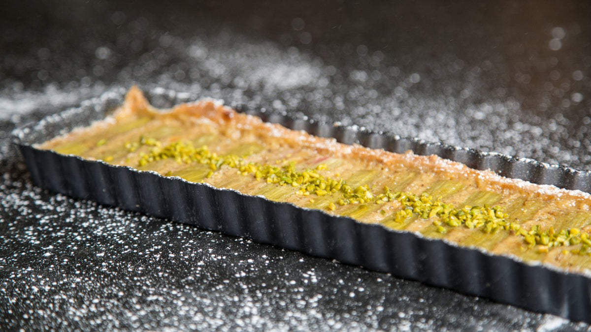 Rechteckige Tarte mit grünem Pistazienbelag in schwarzer Form, bestreut mit gemahlenen Pistazien.